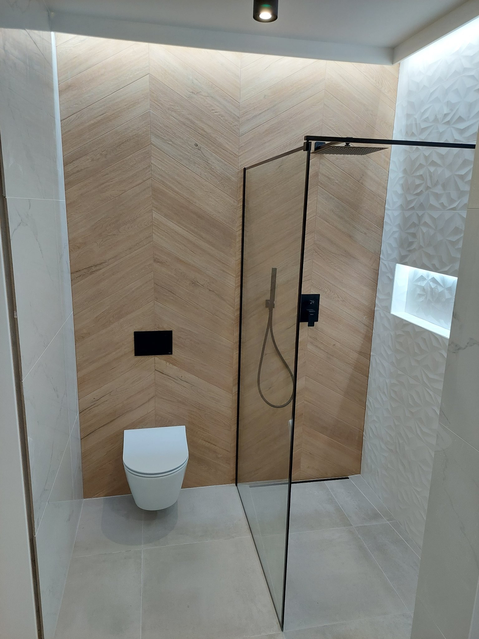 Nowoczesna łazienka z kabiną prysznicową typu walk-in, wiszącą toaletą i drewnopodobnymi płytkami ułożonymi w jodełkę na ścianie, oświetlona punktowo.