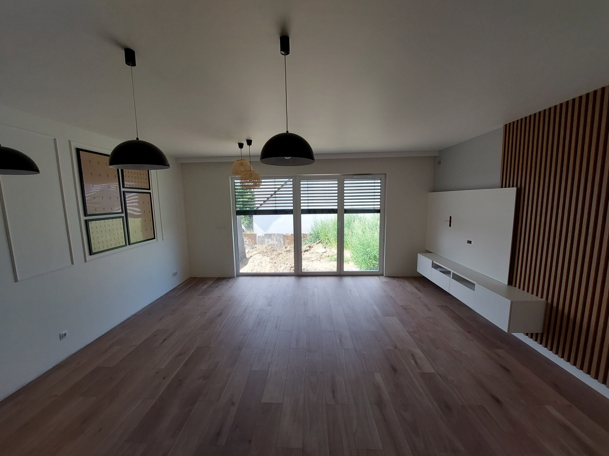 Nowoczesne wnętrze salonu z drewnianą podłogą, białymi ścianami z dekoracyjnymi listwami, drewnianą ścianką działową, wiszącą szafką RTV i trzema lampami wiszącymi w stylu industrialnym.