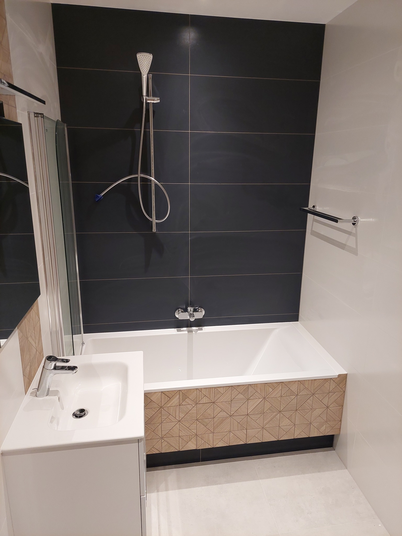 Nowoczesna łazienka z czarnymi płytkami na ścianie, białą wanną obudowaną drewnopodobną mozaiką, białą umywalką z szafką i chromowaną armaturą.