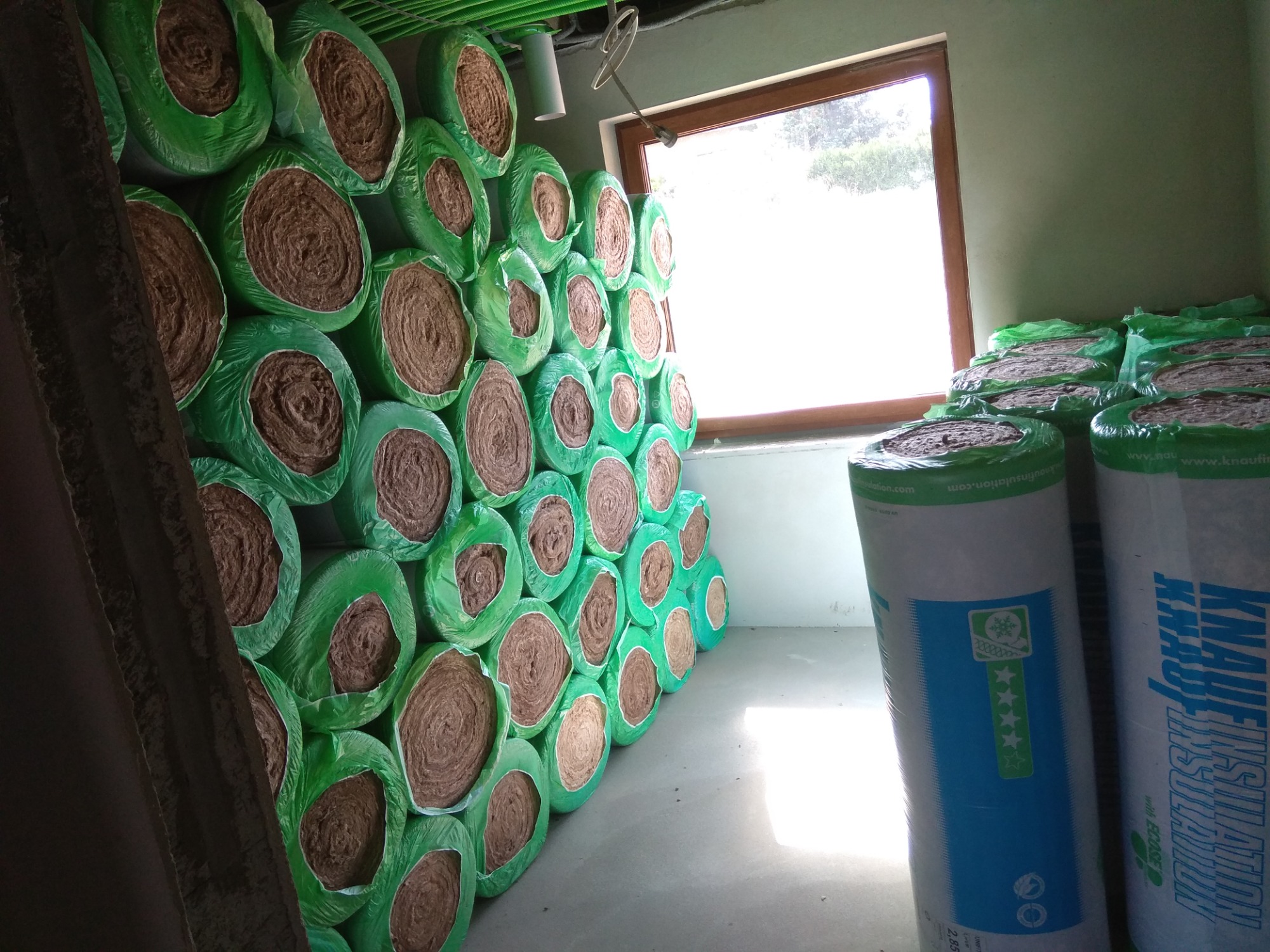 Stos rolek wełny mineralnej w zielonym opakowaniu, ułożonych przy ścianie w pomieszczeniu z oknem, widoczne etykiety producenta Knauf Insulation.