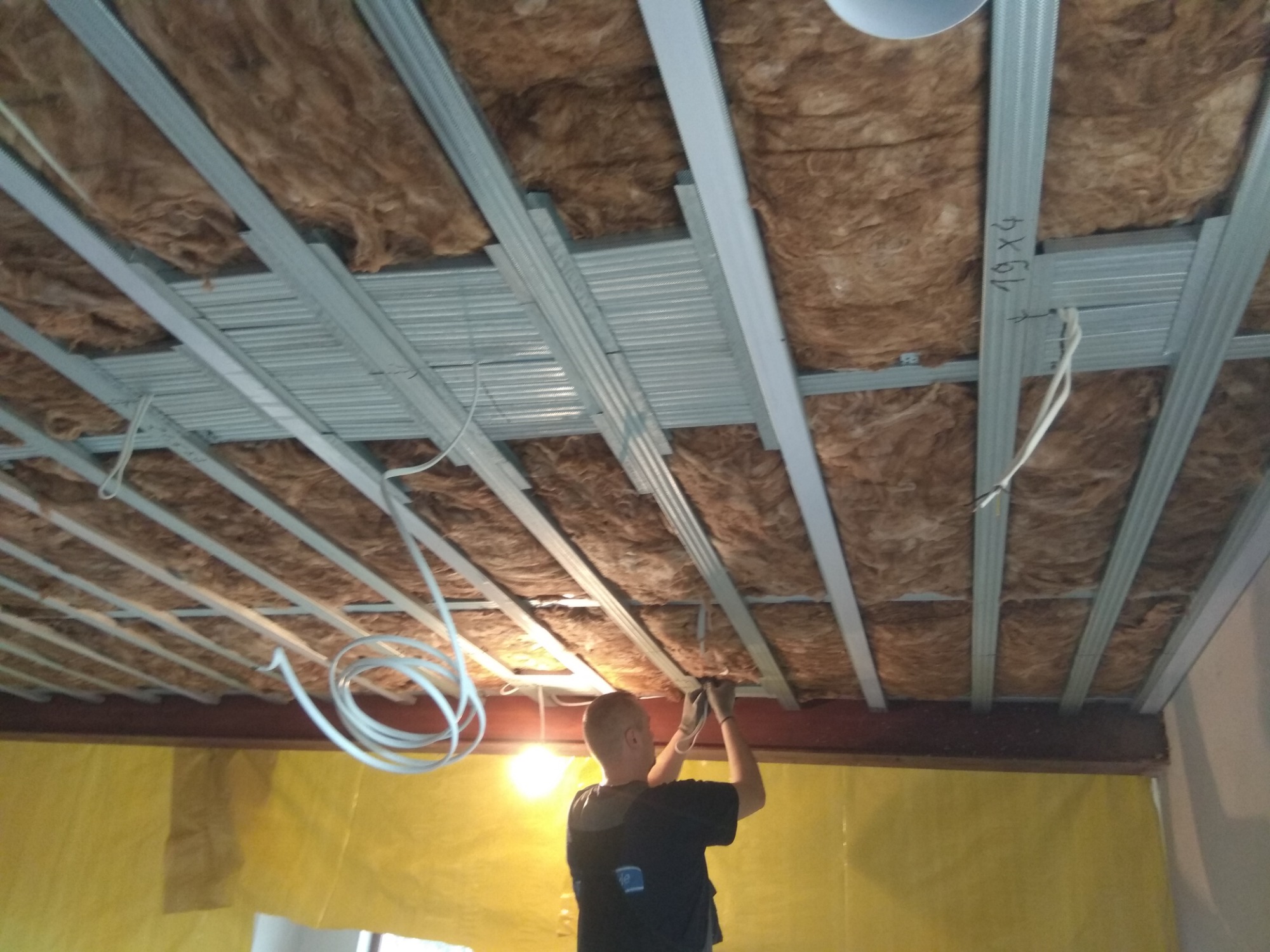 Monteraż sufitu podwieszanego z izolacją z wełny mineralnej, widoczne profile metalowe i instalacja elektryczna, pracownik w trakcie montażu.
