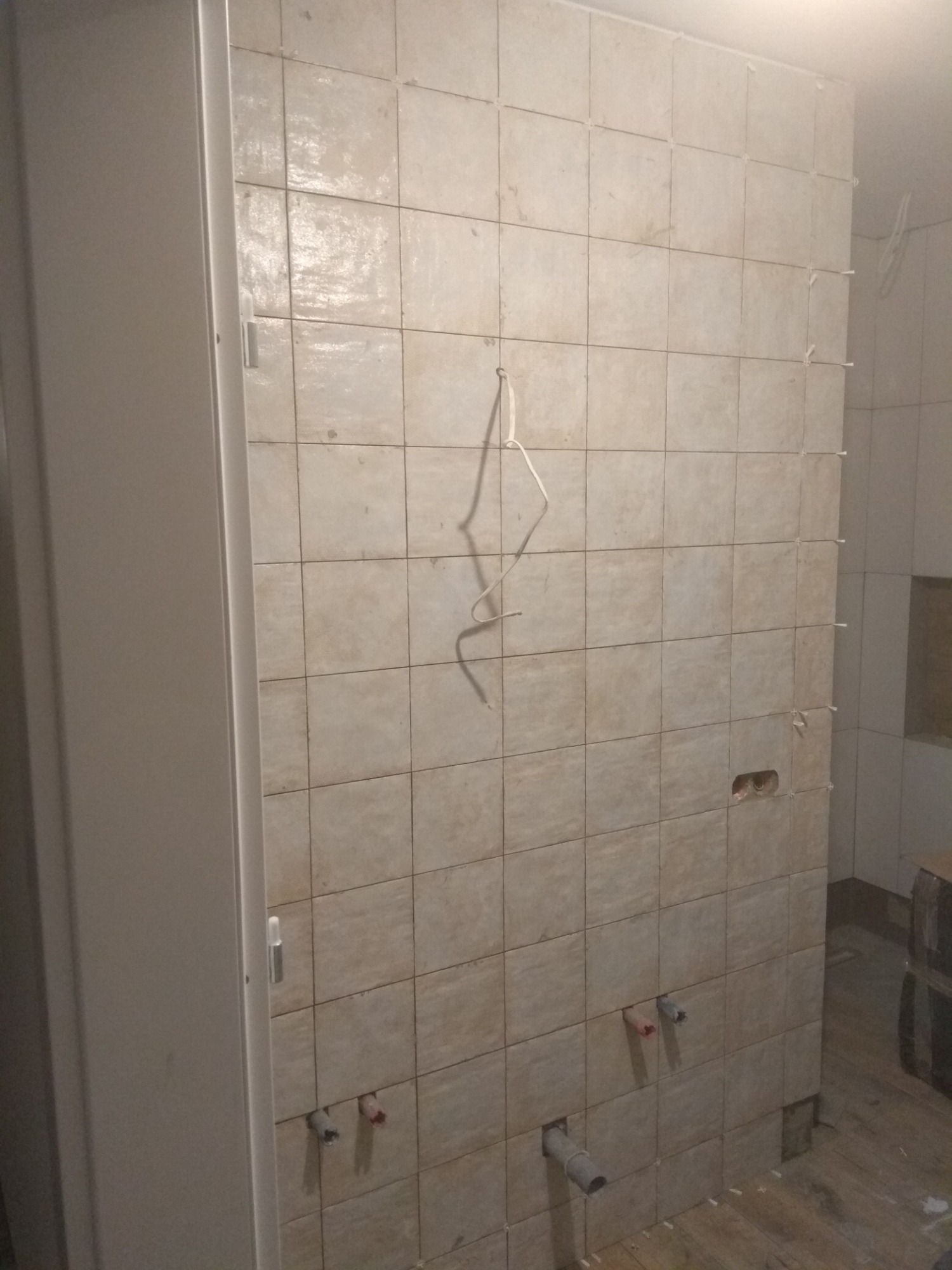 Ściana w trakcie układania płytek ceramicznych, widoczne krzyżyki dystansowe, wystające rury instalacji wodnej i fragment białej ościeżnicy drzwiowej.