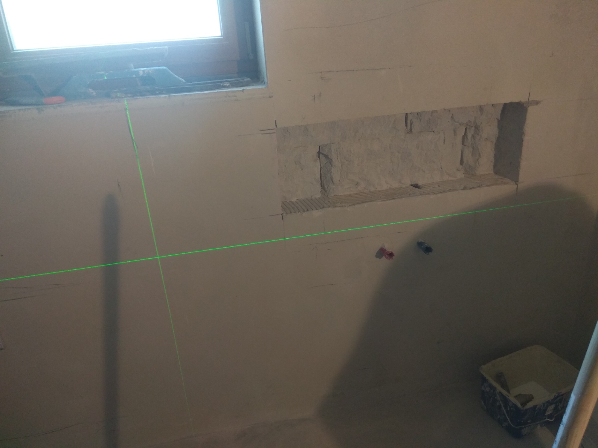 Przygotowanie ściany pod glazurę: wycięty otwór na półkę, widoczne ślady po ołówku, poziomica laserowa i pojemnik z zaprawą.