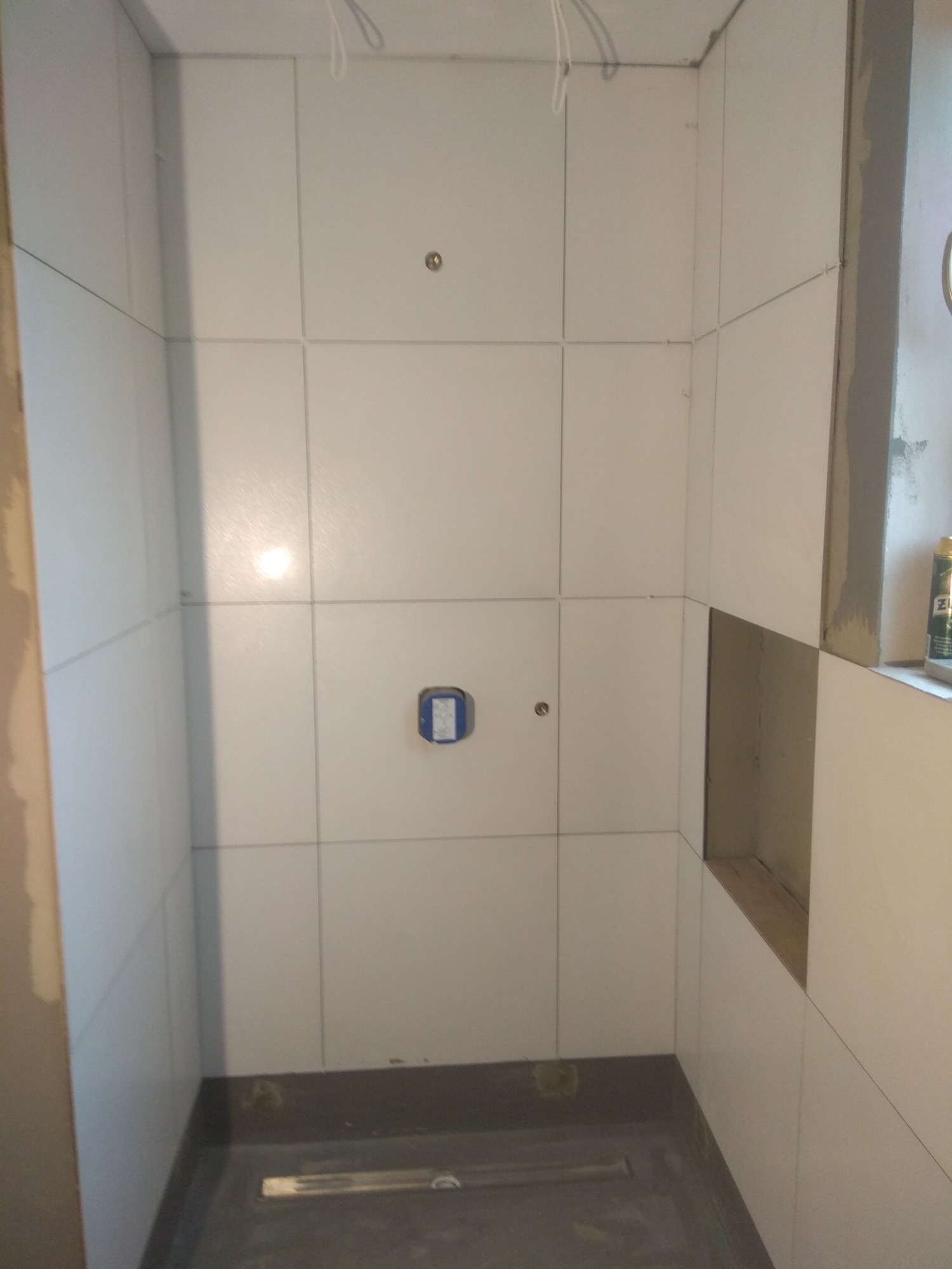 Wnętrze kabiny prysznicowej w trakcie wykańczania, widoczne białe płytki ułożone na ścianach, otwór w ścianie przygotowany pod półkę, zainstalowany odpływ liniowy w podłodze, puszka elektryczna...