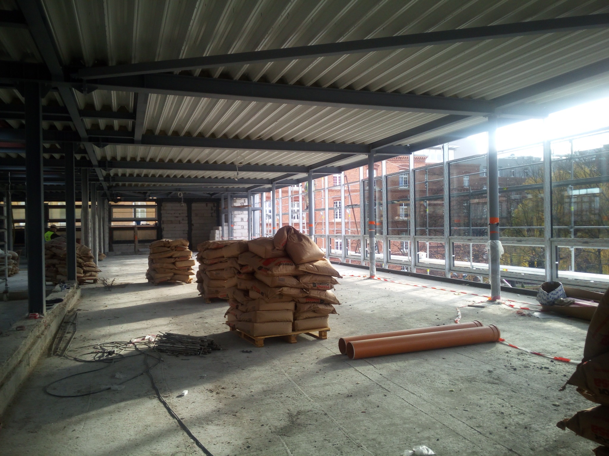 Wnętrze budynku w trakcie budowy z widoczną stalową konstrukcją stropu, paletami z workami cementu i rurami PCV leżącymi na betonowej posadzce. W tle budynek z cegły i okna z widokiem na zewnątrz.