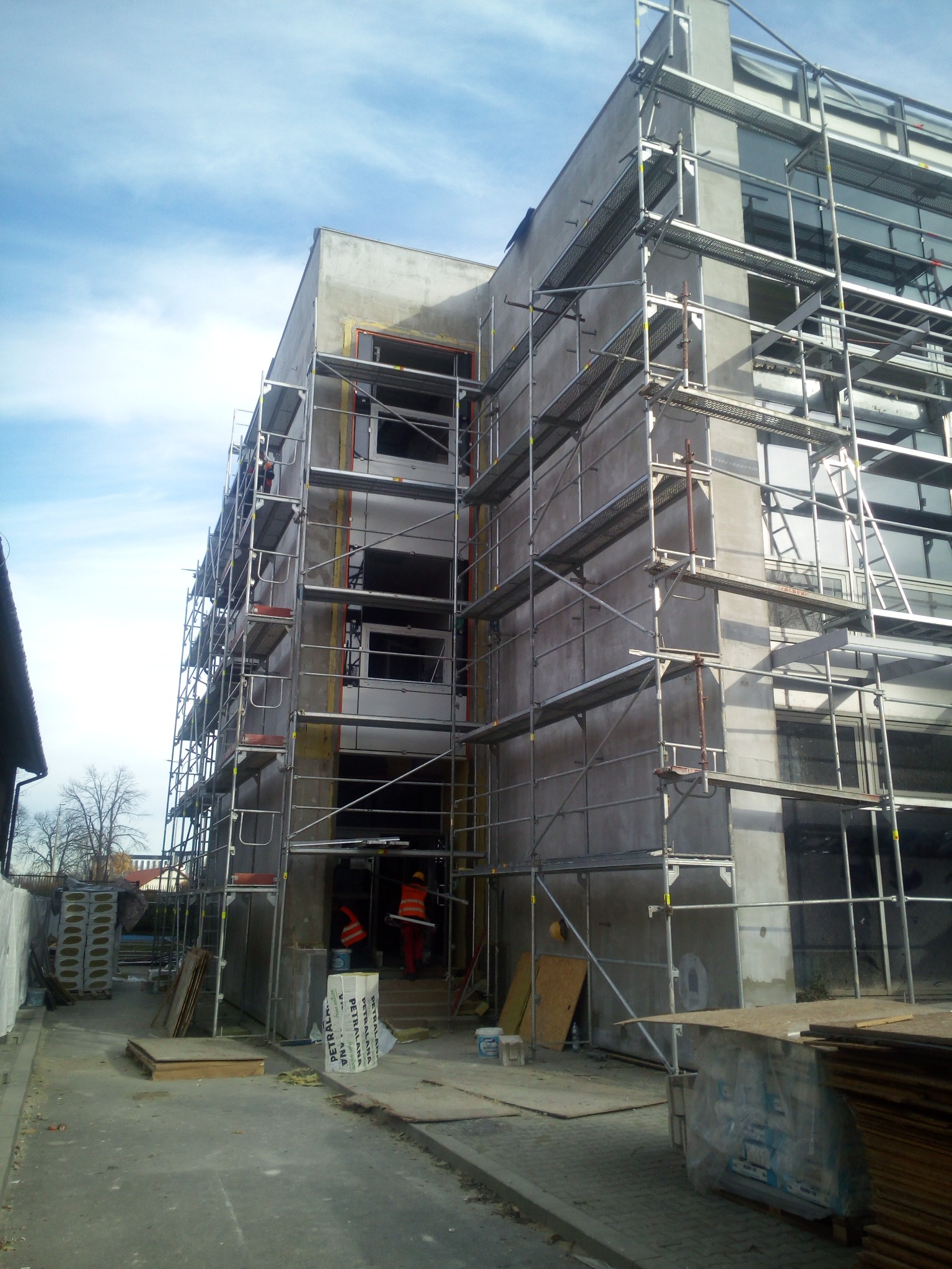 Budynek w trakcie ocieplania, z rusztowaniami, widoczni pracownicy w pomarańczowych kamizelkach, materiały budowlane na placu budowy.