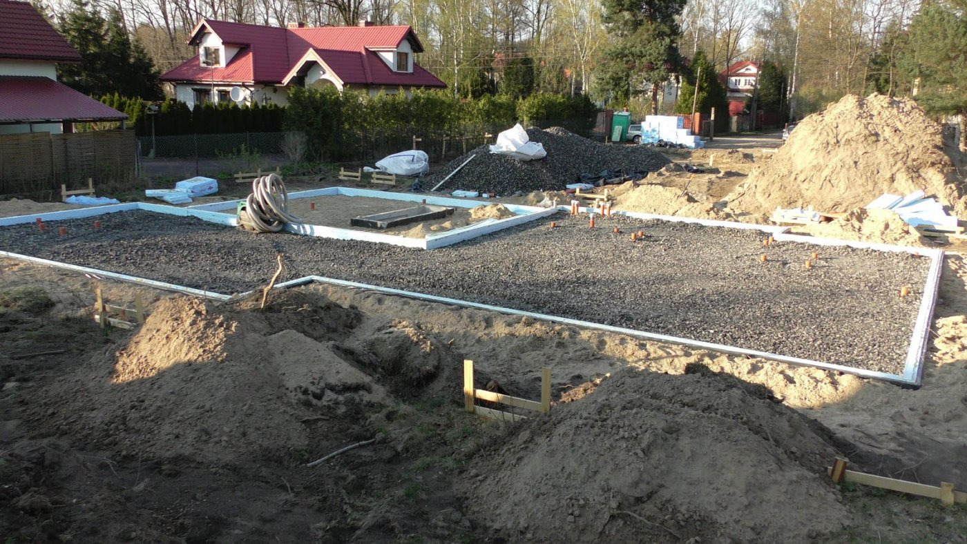 Przygotowany teren pod wylewkę fundamentów: obrys z białych płyt, wysypany żwir, wystające rury kanalizacyjne, w tle domy jednorodzinne.