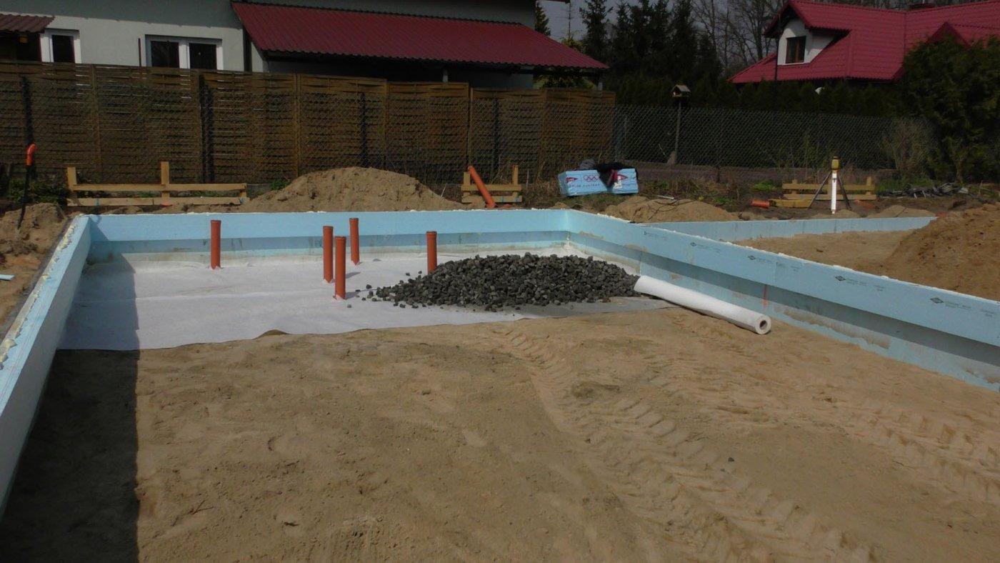 Fundamenty domu jednorodzinnego w trakcie budowy: izolacja ze styropianu, rury kanalizacyjne pionowe, podsypka żwirowa, geowłóknina, widoczne ślady opon na piasku.