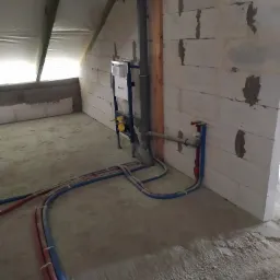 Surowy stan wykończenia łazienki na poddaszu: widoczne rury instalacji wodno-kanalizacyjnej, stelaż podtynkowy WC Geberit i fragment dachu ze skosem.