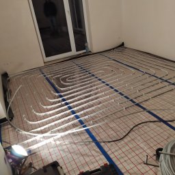 Sjk-Instal Krzysztof Szczepan - Montaż ogrzewania podłogowego: białe rurki ułożone spiralnie na czerwono-białej siatce, niebieskie taśmy mocujące, narzędzia w tle, widoczna osoba w odbiciu szyby, datownik w prawym dolnym rogu.