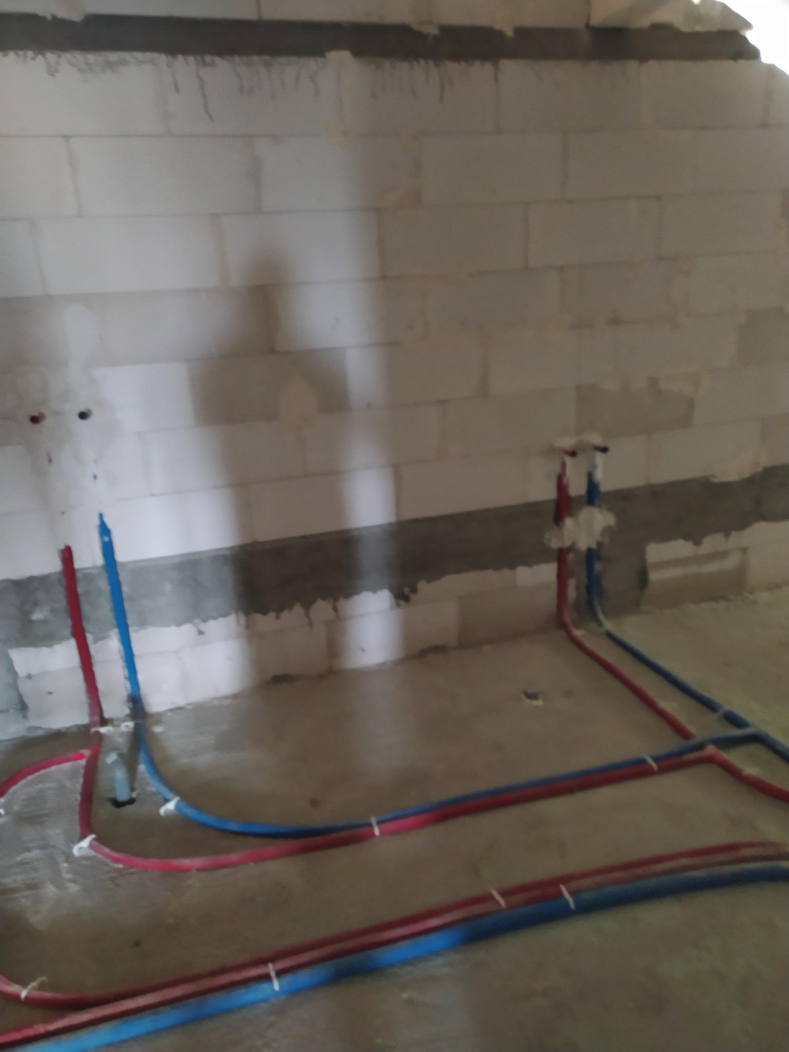 Instalacja rur wodnych i grzewczych w surowym stanie, czerwone i niebieskie rury prowadzone po podłodze w pomieszczeniu z murem z betonu komórkowego.