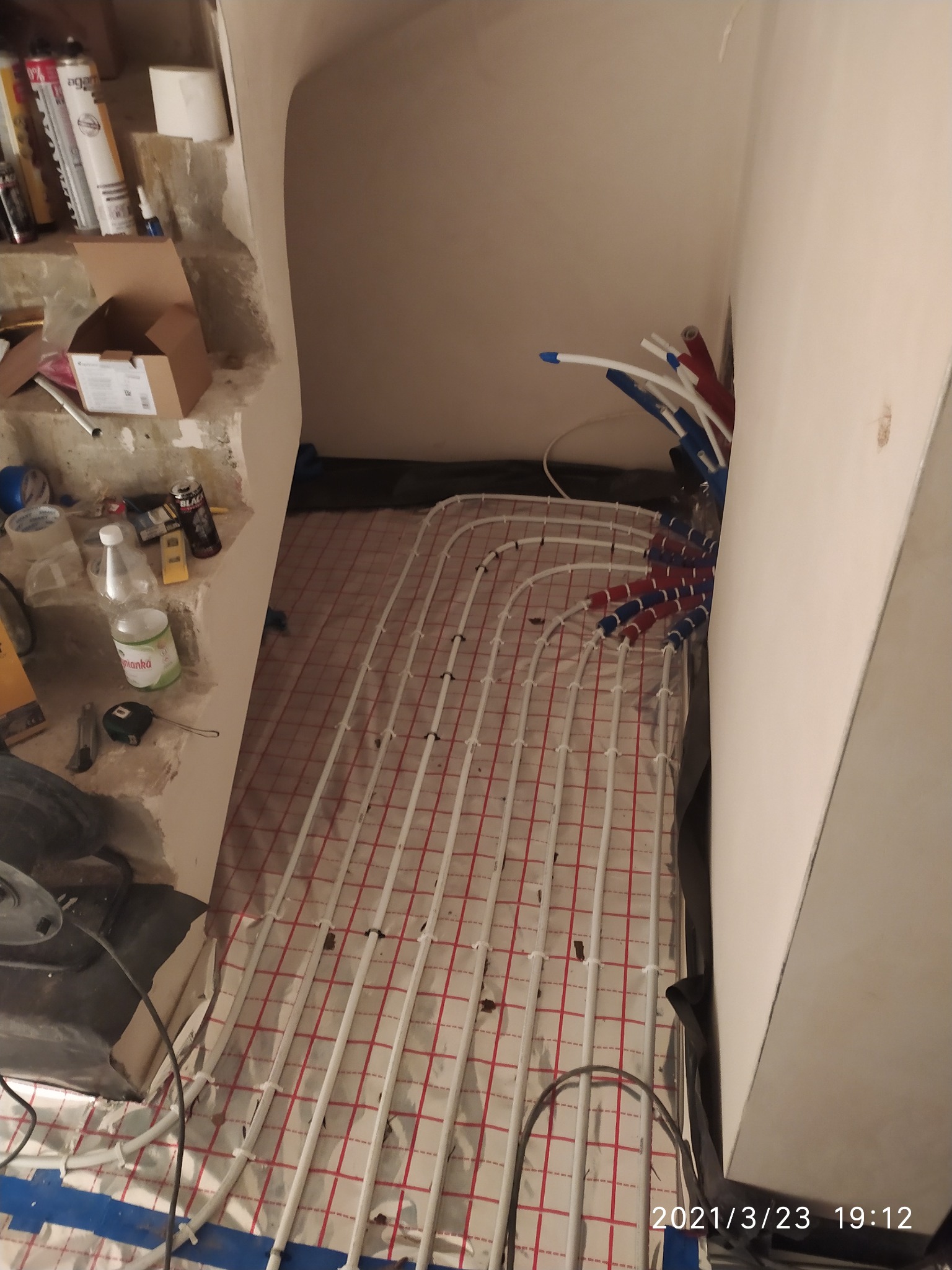 Rury ogrzewania podłogowego ułożone na folii z siatką, widoczne narzędzia pracy i materiały budowlane na nierównym podłożu, przygotowania do wylewki.