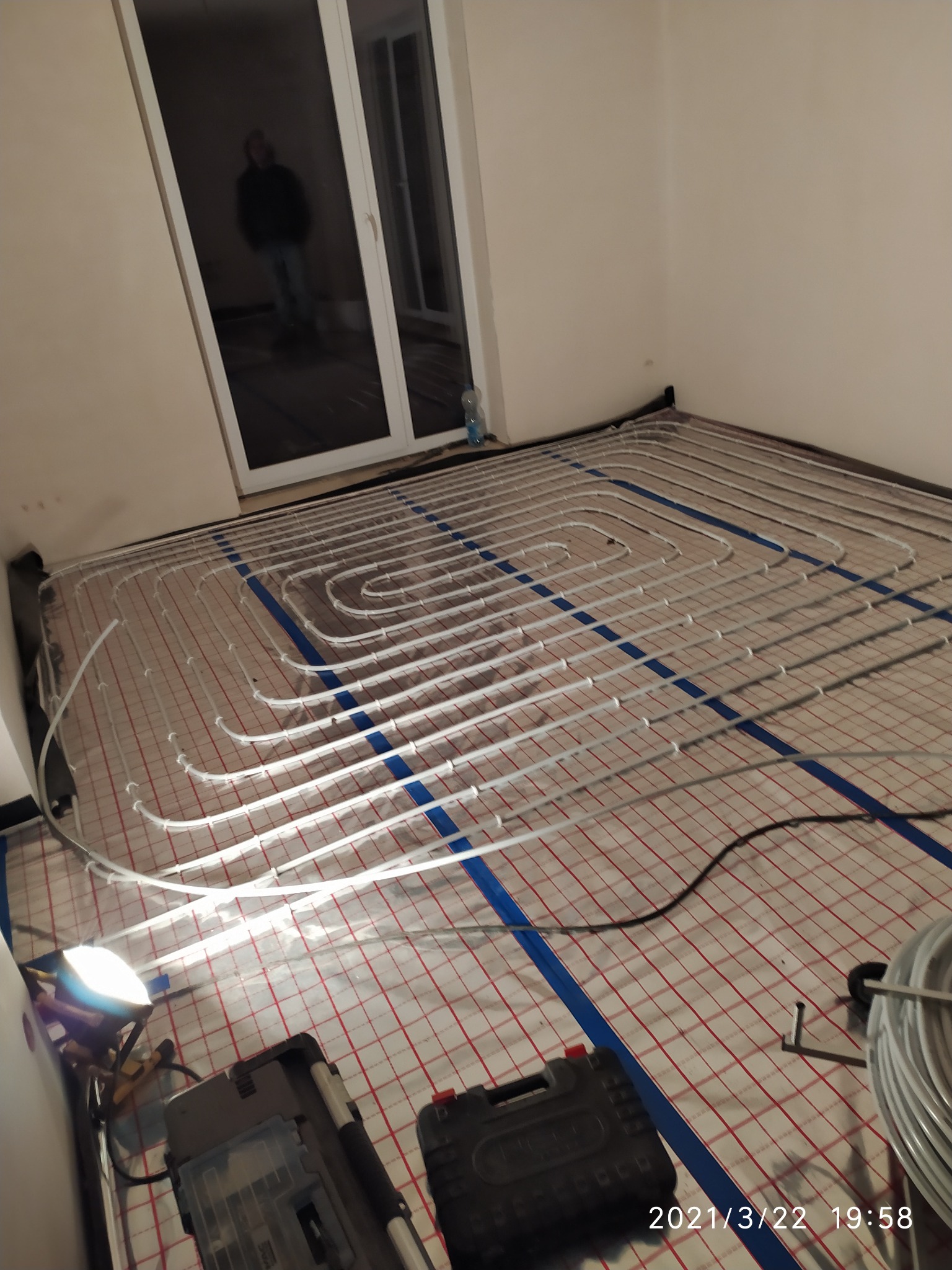 Montaż ogrzewania podłogowego: białe rurki ułożone spiralnie na czerwono-białej siatce, niebieskie taśmy mocujące, narzędzia w tle, widoczna osoba w odbiciu szyby, datownik w prawym dolnym rogu.