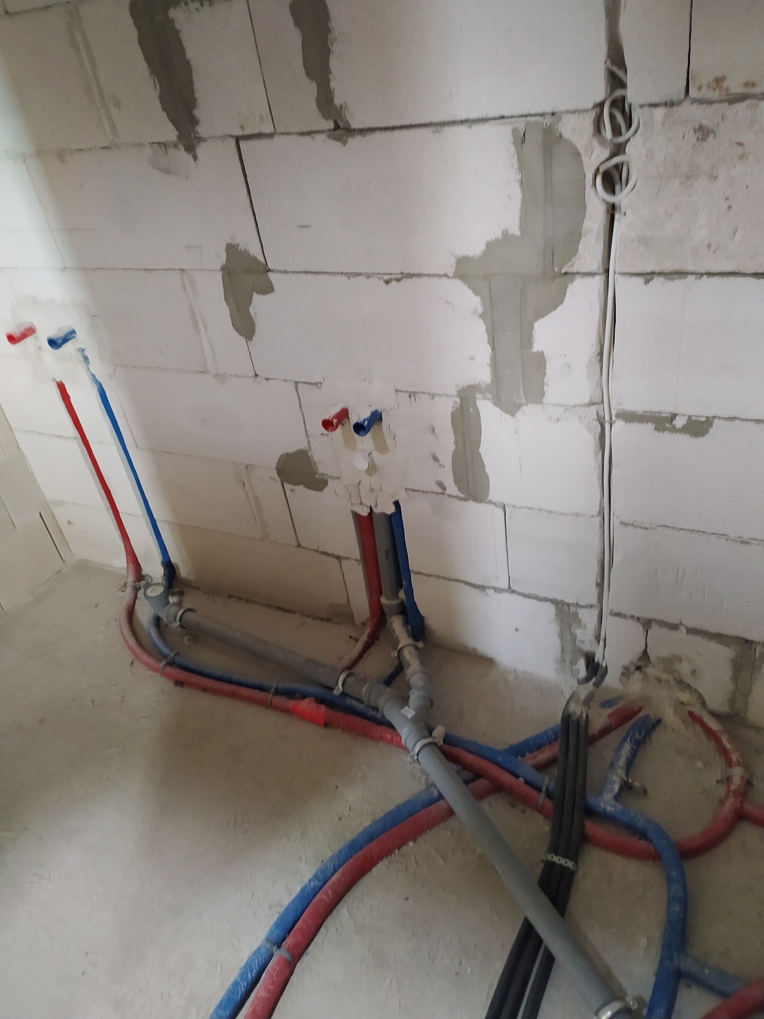 Wewnętrzna instalacja wodna i grzewcza w surowym stanie, rury z tworzywa sztucznego w kolorach czerwonym i niebieskim prowadzone po podłodze i wyprowadzone ze ściany z betonu komórkowego, widoczne...