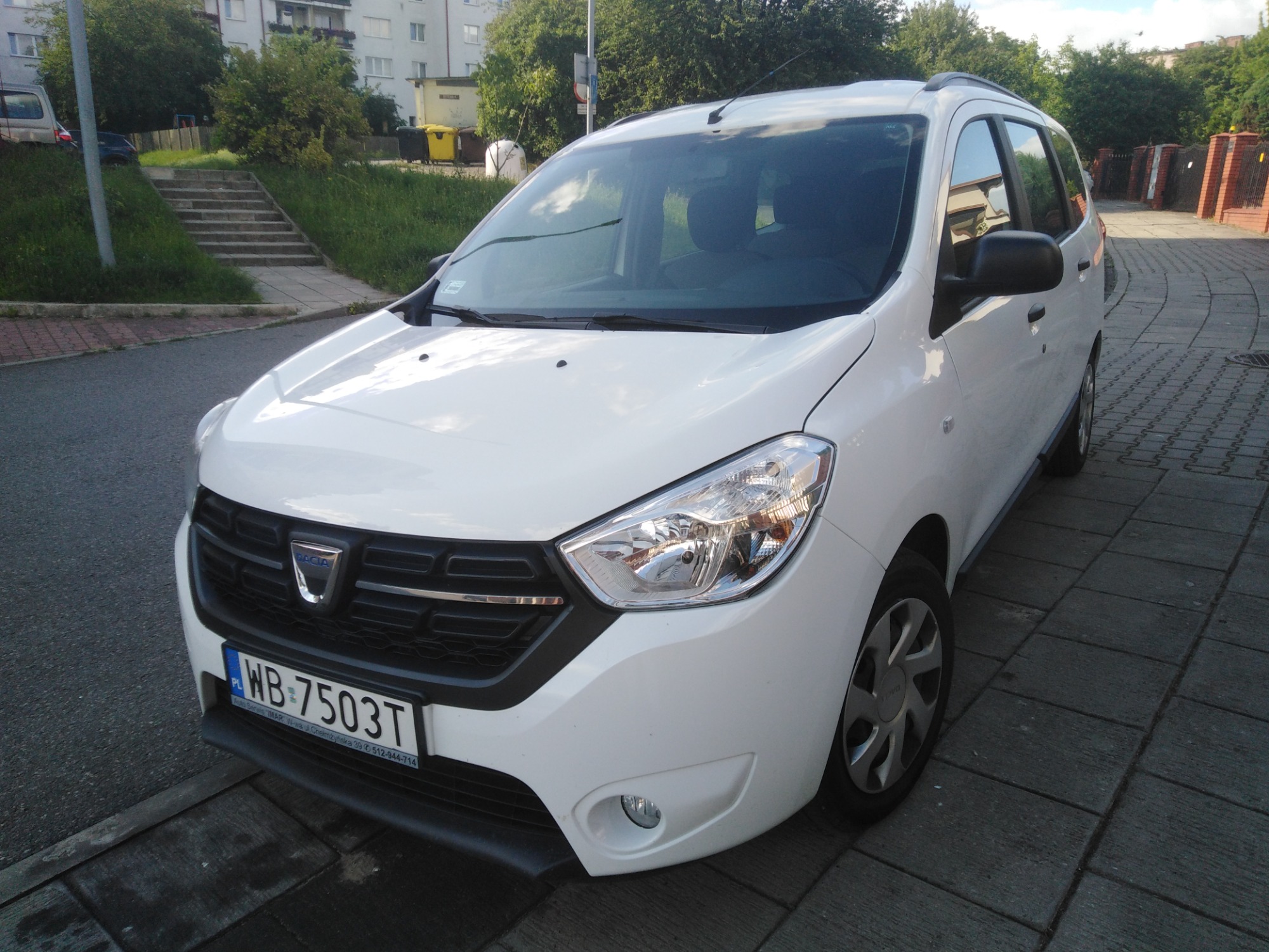 Biały samochód typu kombi Dacia Lodgy Stepway zaparkowany na chodniku z szarej kostki brukowej, widok z przodu z lekkiego ukosa, widoczna tablica rejestracyjna WB-7503T.
