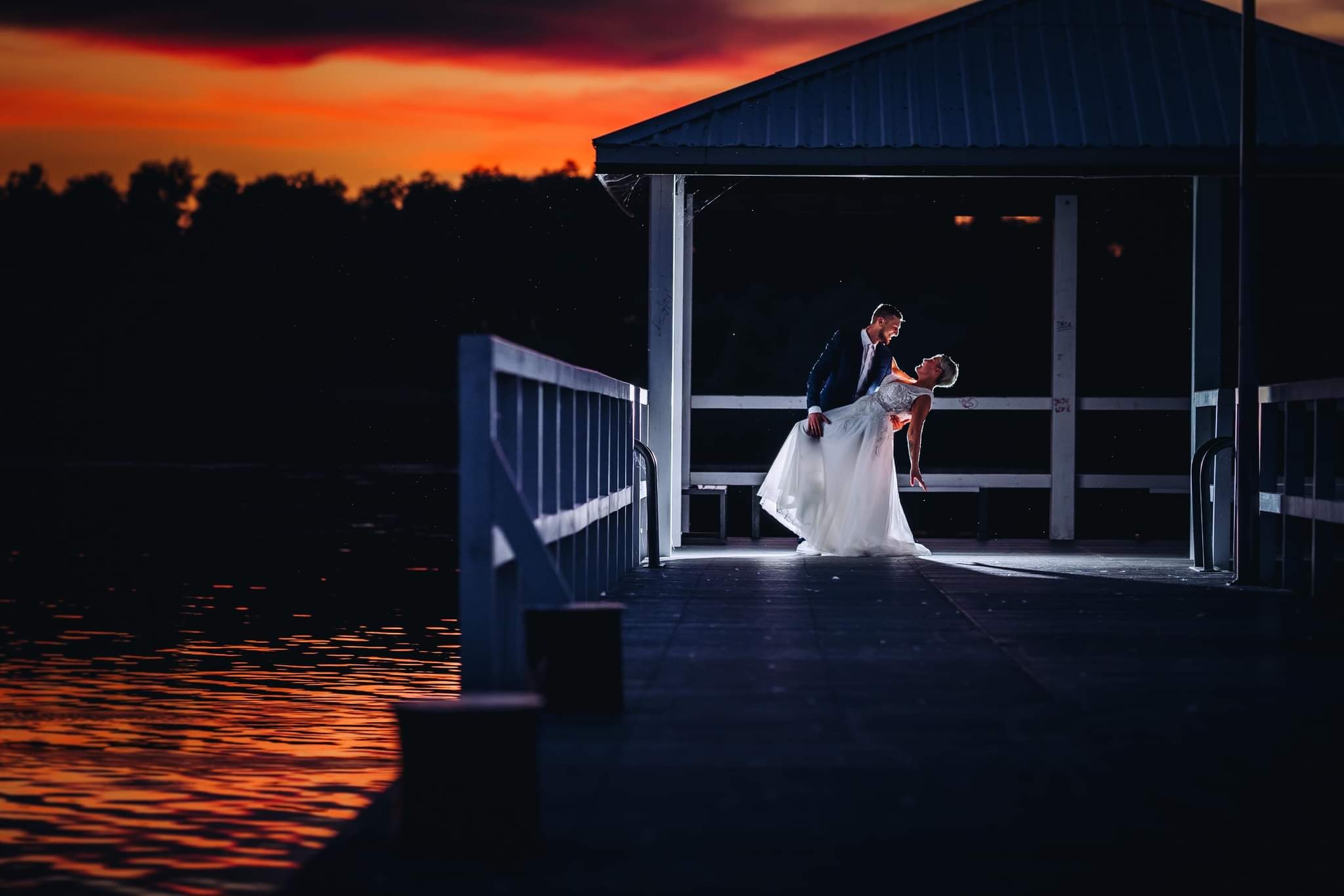 Para młoda tańczy na drewnianym pomoście nad wodą o zachodzie słońca, oświetlona światłem z altany.