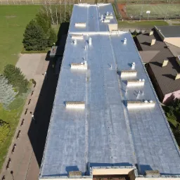 Srebrzysty dach przemysłowy z licznymi kominkami wentylacyjnymi, widok z góry, otoczony zielenią i budynkami. Blacha trapezowa na dachu, kominki wentylacyjne.