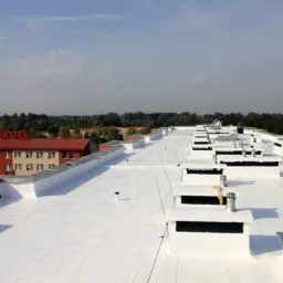 Biały, płaski dach budynku komercyjnego po renowacji, widok z góry, na tle hotel z czerwonym napisem, w oddali drzewa i błękitne niebo.
