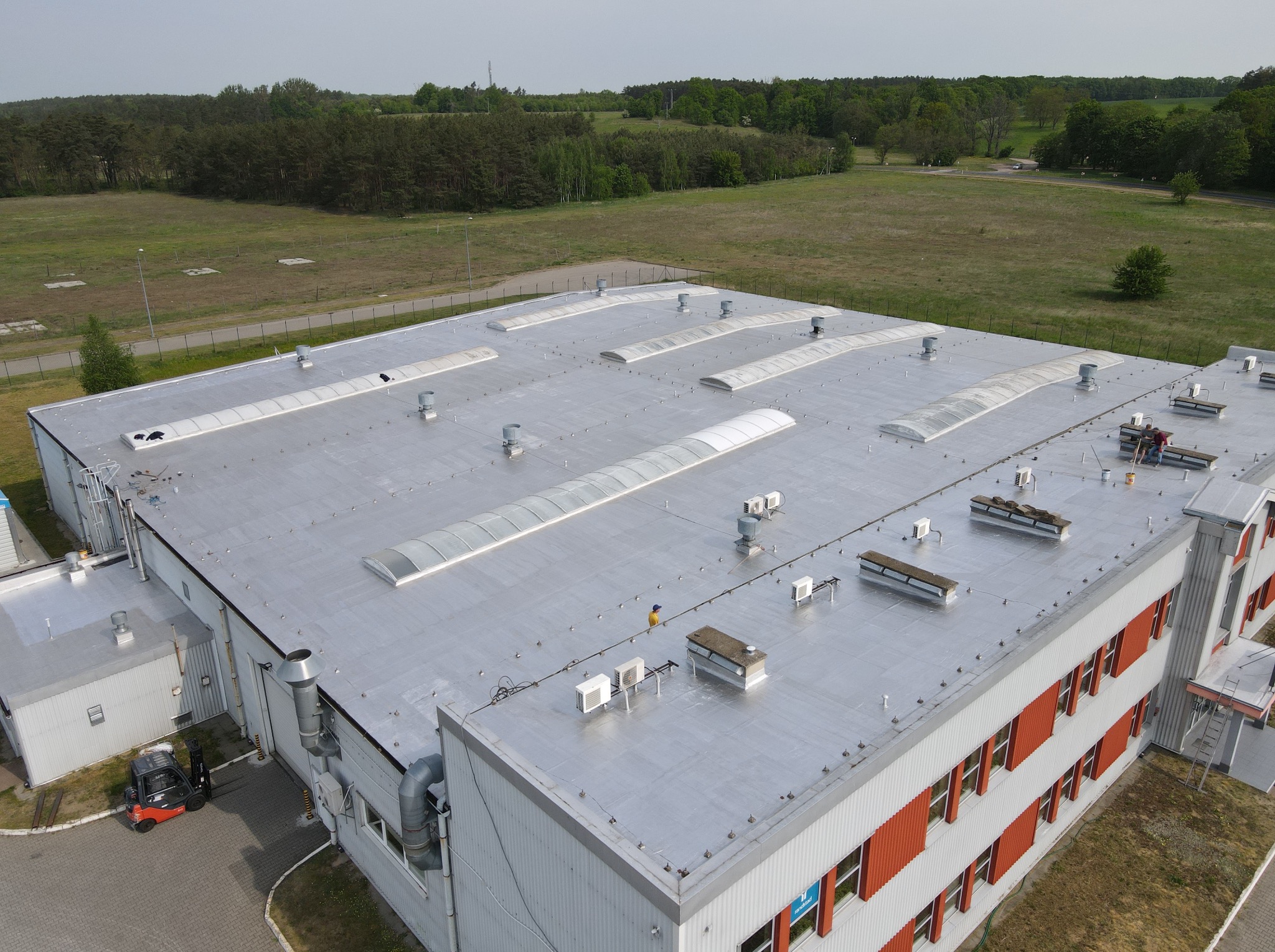 Srebrny dach przemysłowy z świetlikami i wentylacją, widok z góry. Na dachu robotnicy i klimatyzatory. W tle las i łąka, na dole wózek widłowy.