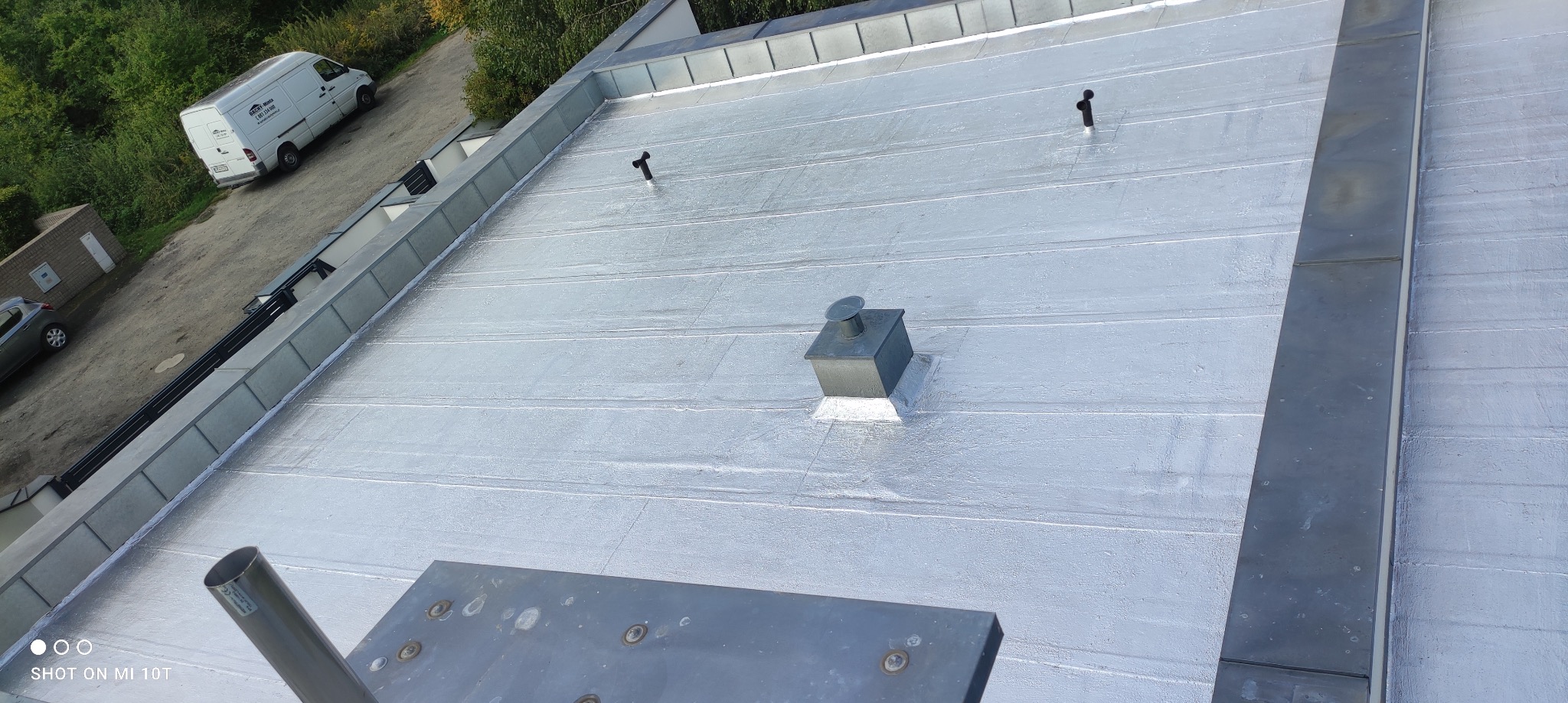 Wykończony dach z blachy na rąbek stojący, widok z góry. Na dachu komin wentylacyjny i odpowietrzniki. W tle budynek i samochód dostawczy firmy dekarskiej.