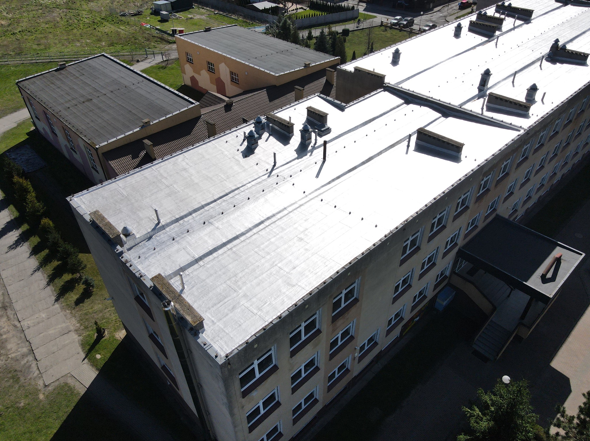 Jasnoszary dach budynku wielopiętrowego widziany z góry, z widocznymi kominami i systemem wentylacyjnym. Nowe pokrycie dachu, bez uszkodzeń.