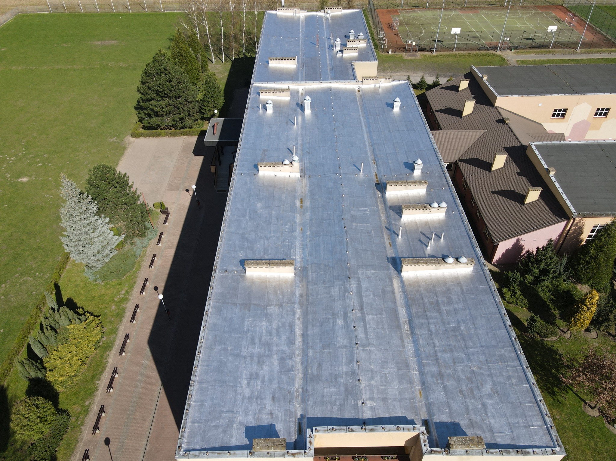 Srebrzysty dach przemysłowy z licznymi kominkami wentylacyjnymi, widok z góry, otoczony zielenią i budynkami. Blacha trapezowa na dachu, kominki wentylacyjne.