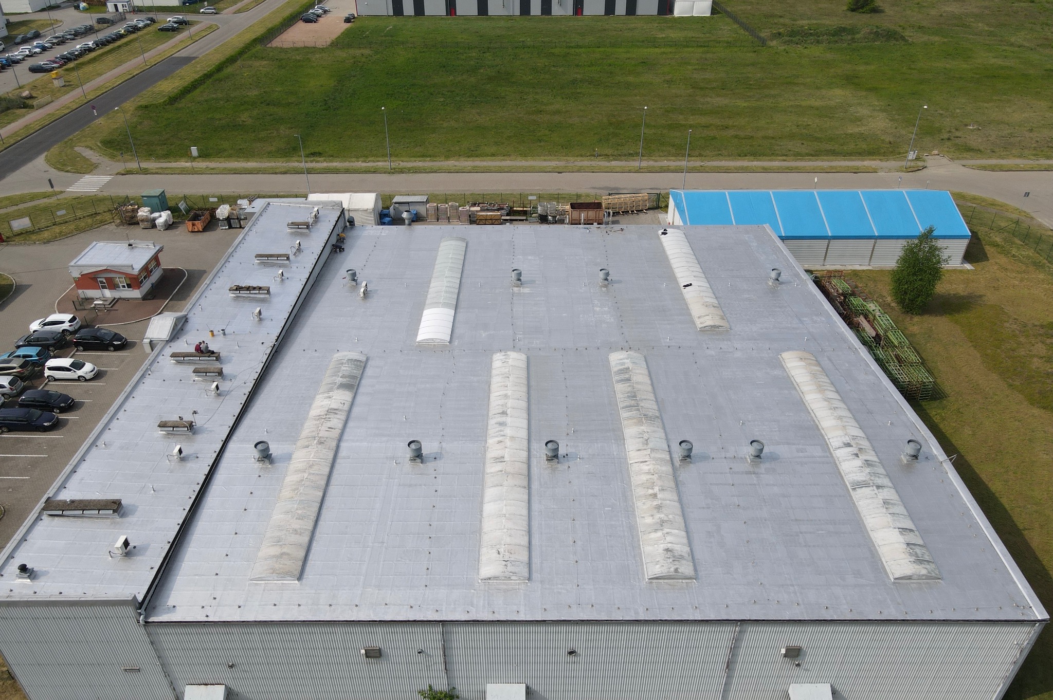 Szeroki, płaski dach przemysłowy z świetlikami i wentylacją, widok z góry. W tle budynki, trawnik i parking z samochodami.