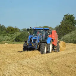 Niebieski traktor z czerwoną prasą zwijającą słomę na polu, w tle widoczne drzewa i okrągłe bele słomy.