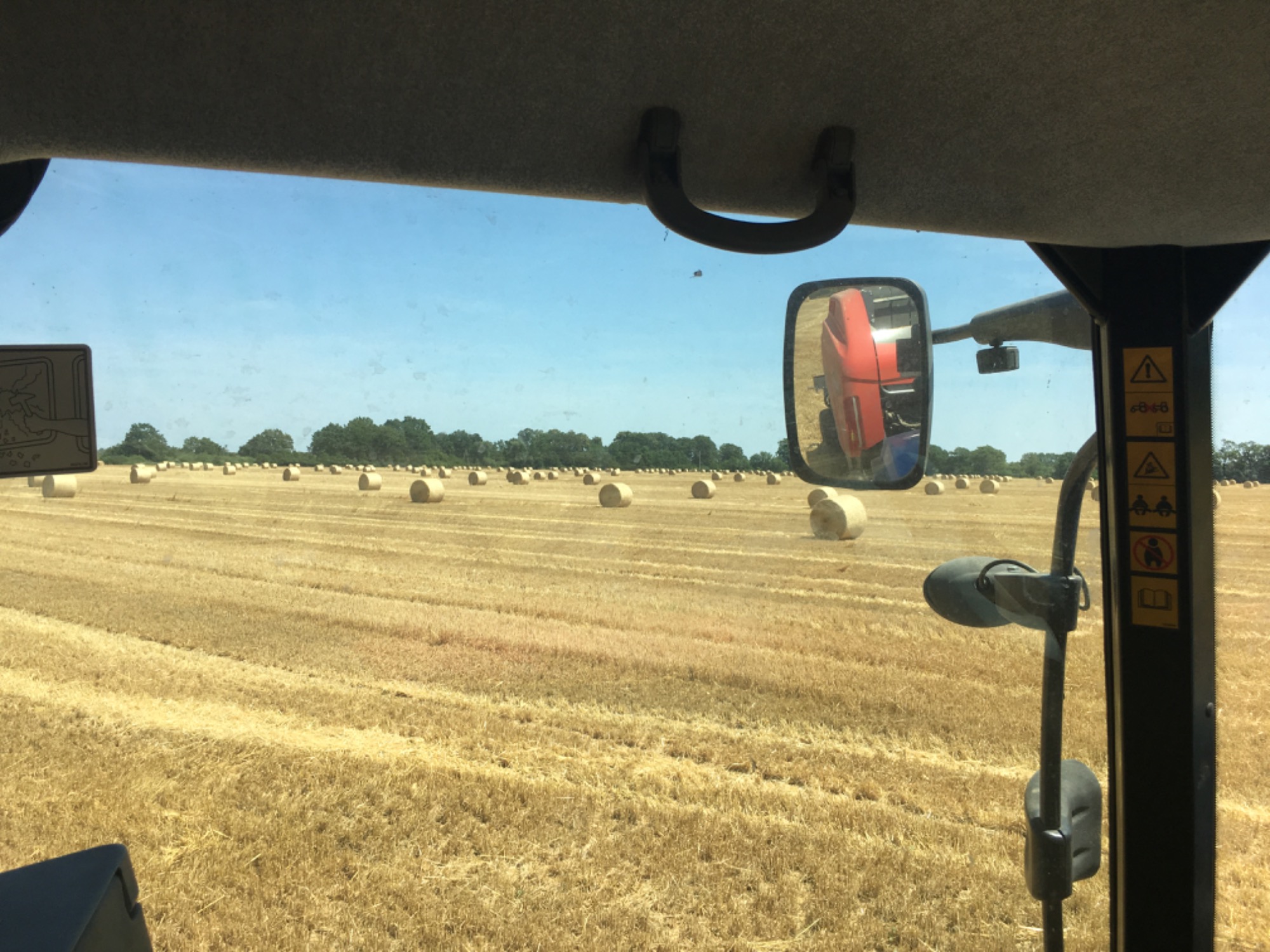 Widok z kabiny traktora na pole ze słomą i okrągłymi balotami, z lusterka wstecznego widać kombajn.