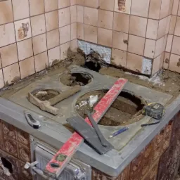 Fragment rozebranego pieca kaflowego w rogu pomieszczenia z odspojonymi płytkami, widoczne narzędzia murarskie i szczelina w ścianie.