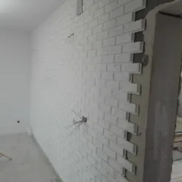 Ściana wyłożona białymi płytkami imitującymi cegłę, z widocznymi kablami elektrycznymi wystającymi ze ściany i niedokończonym narożnikiem przy ościeżnicy.