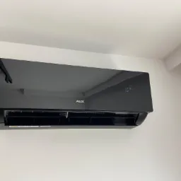 Czarny klimatyzator AUX J-Smart Art zamontowany na białej ścianie, wyświetlający temperaturę 16 stopni Celsjusza.