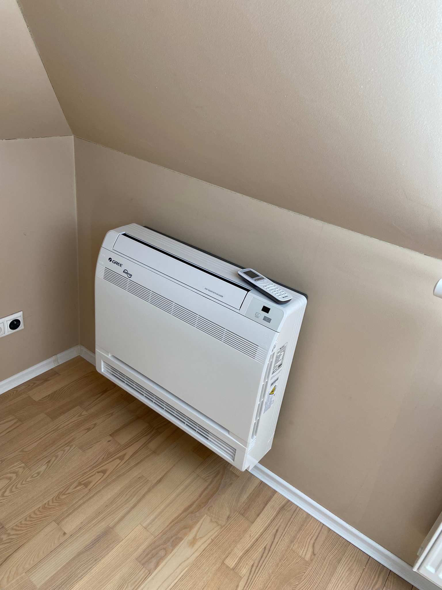 Biały klimatyzator podłogowy Gree z pilotem na górze, zamontowany w pokoju na poddaszu z drewnianą podłogą i beżowymi ścianami.