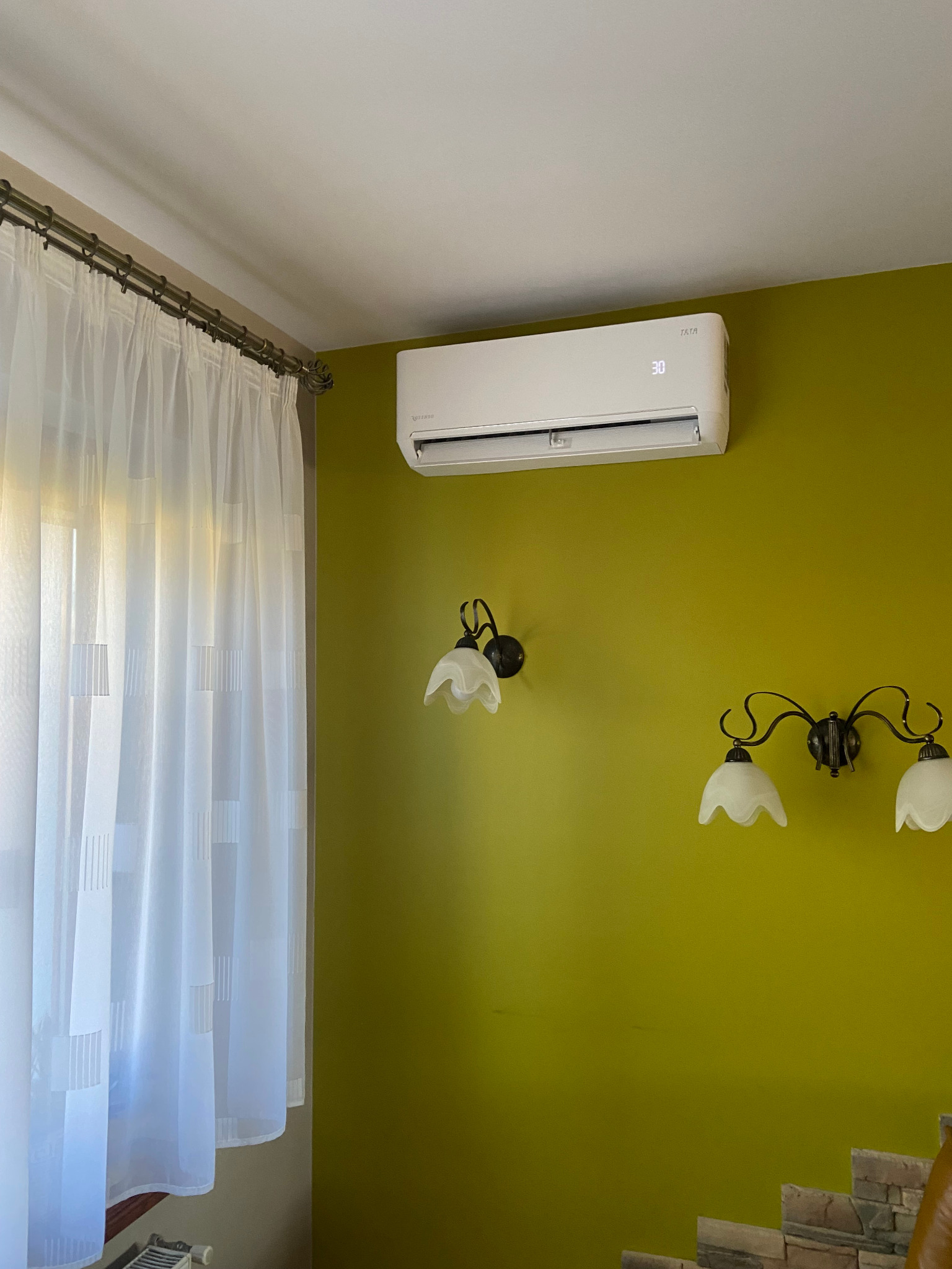 Zamontowany biały klimatyzator na zielonej ścianie w pomieszczeniu w Biadaczu, obok okna z białymi zasłonami i ozdobnych kinkietów.