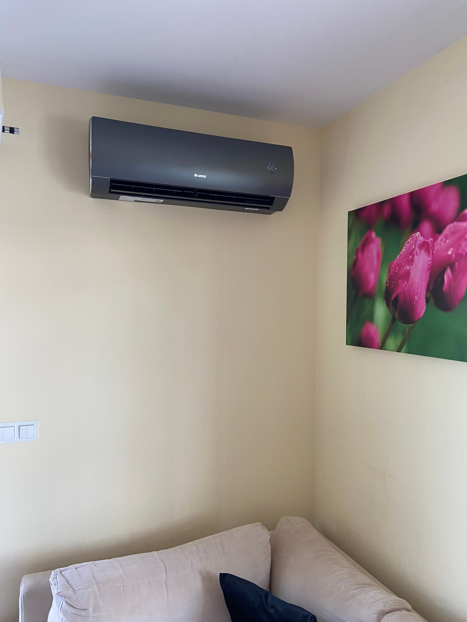 Szary klimatyzator marki Gree zamontowany na kremowej ścianie nad beżową sofą z granatową poduszką, obok wisi obraz z różowymi tulipanami.
