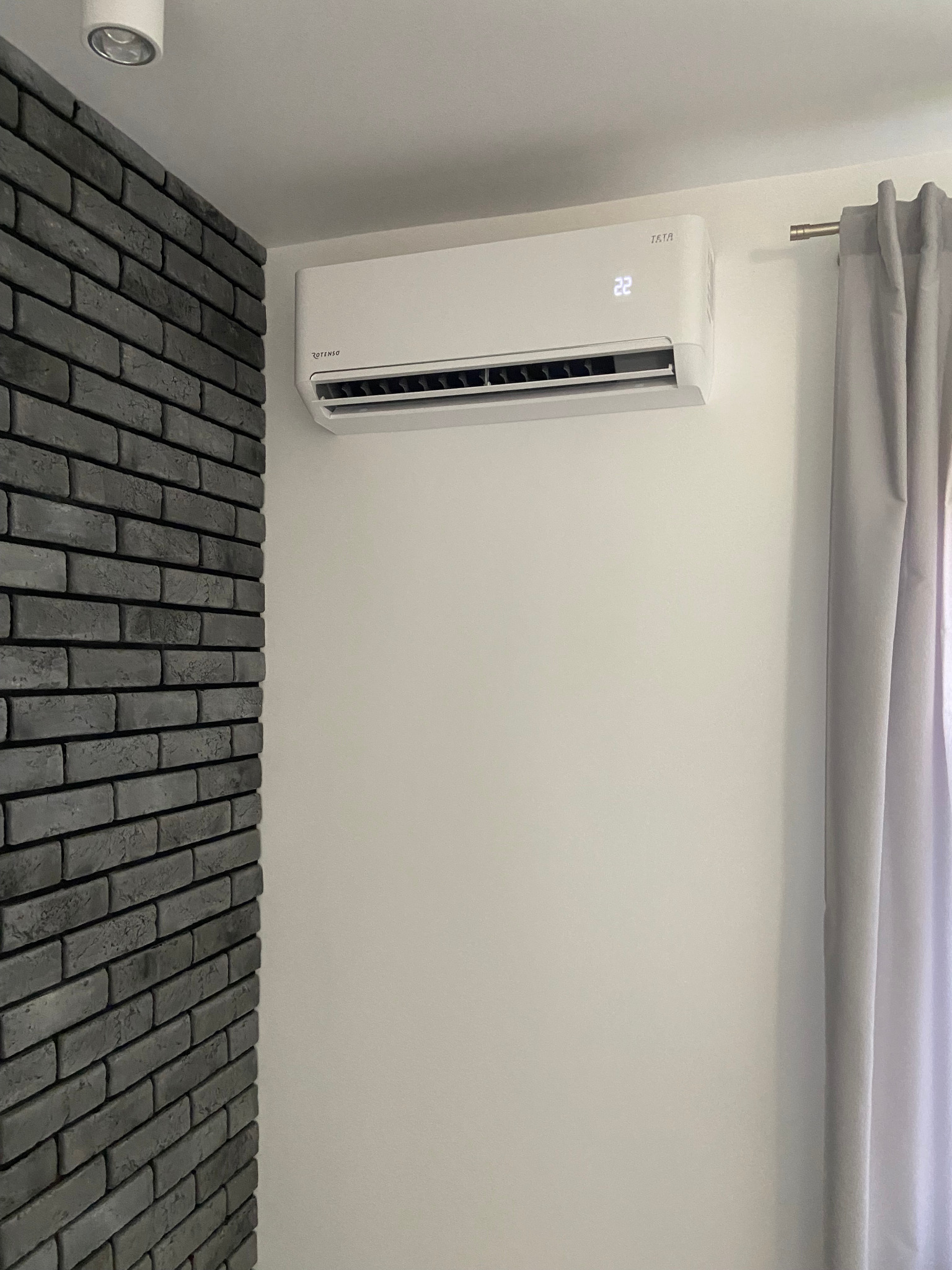 Biały klimatyzator marki Rotenso zamontowany na białej ścianie, obok szarej ściany z cegieł i zasłony. Wyświetlacz pokazuje temperaturę 22 stopnie.