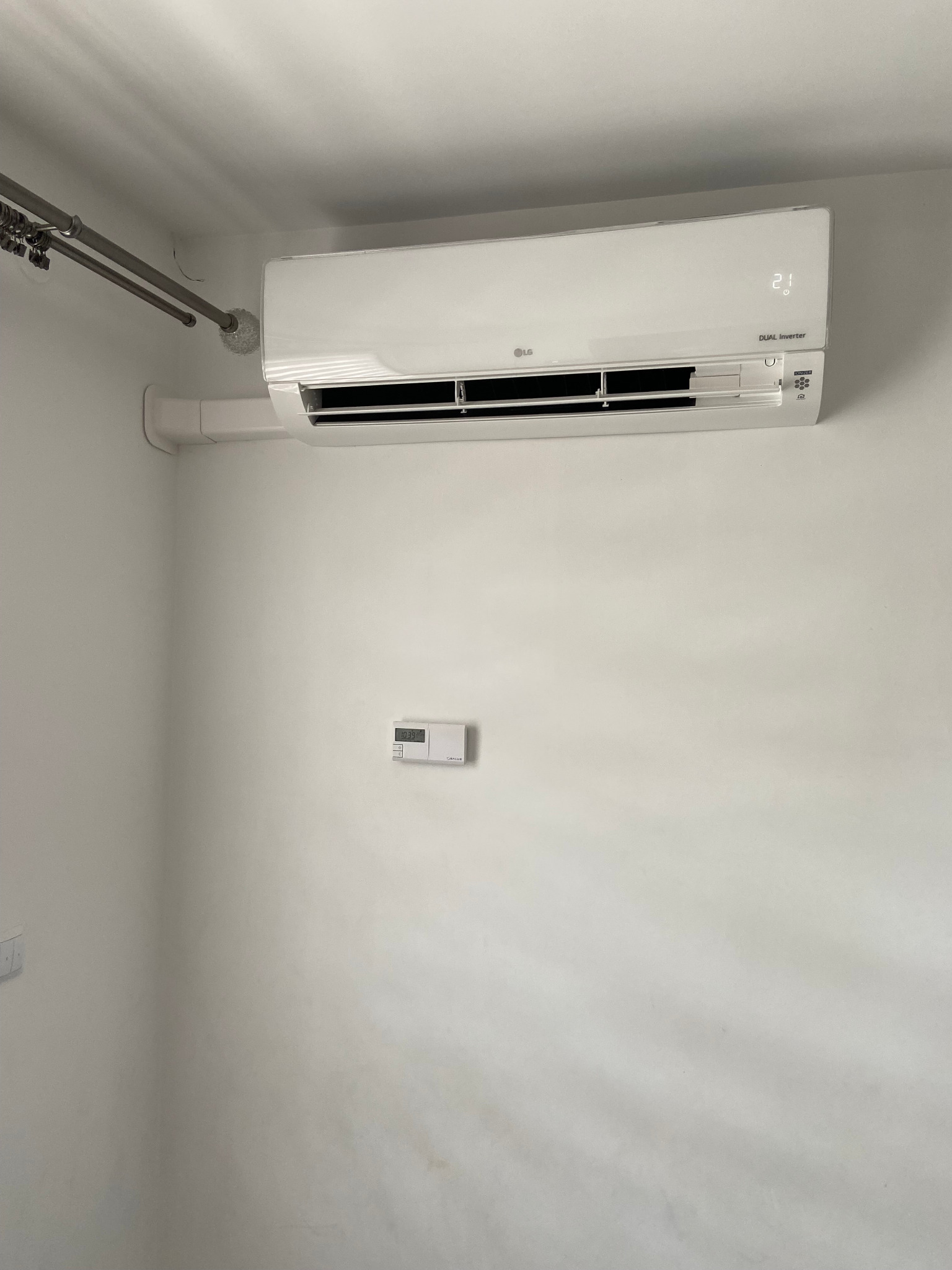 Biały klimatyzator LG Dual Inverter zamontowany na białej ścianie, z widocznym termostatem na ścianie poniżej i rurami instalacyjnymi w górnym rogu.