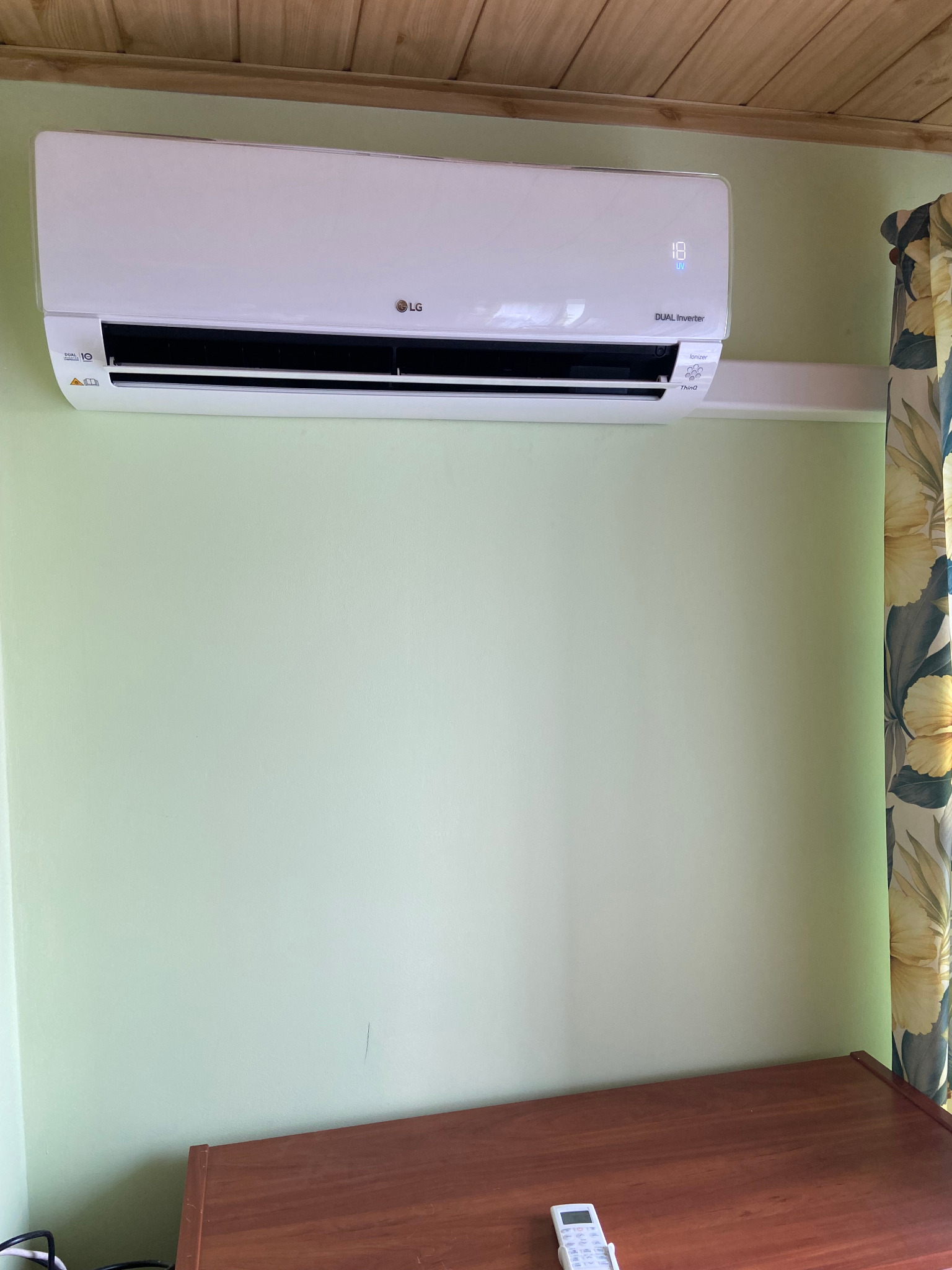 Biały klimatyzator LG Dual Inverter zamontowany na zielonej ścianie w pomieszczeniu z drewnianym sufitem i kwiatowymi zasłonami, z pilotem leżącym na drewnianej komodzie poniżej.