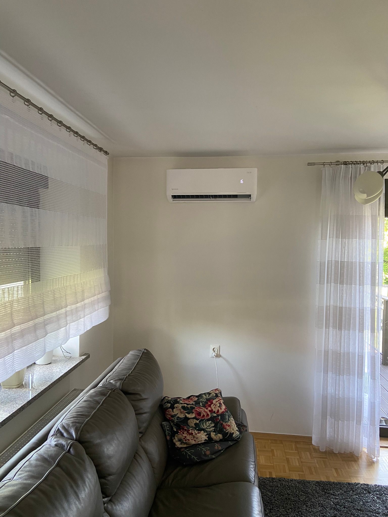 Biały klimatyzator ścienny TATA zamontowany w jasnym salonie z szarym skórzanym narożnikiem i wzorzystą poduszką, widoczne firanki w oknach.