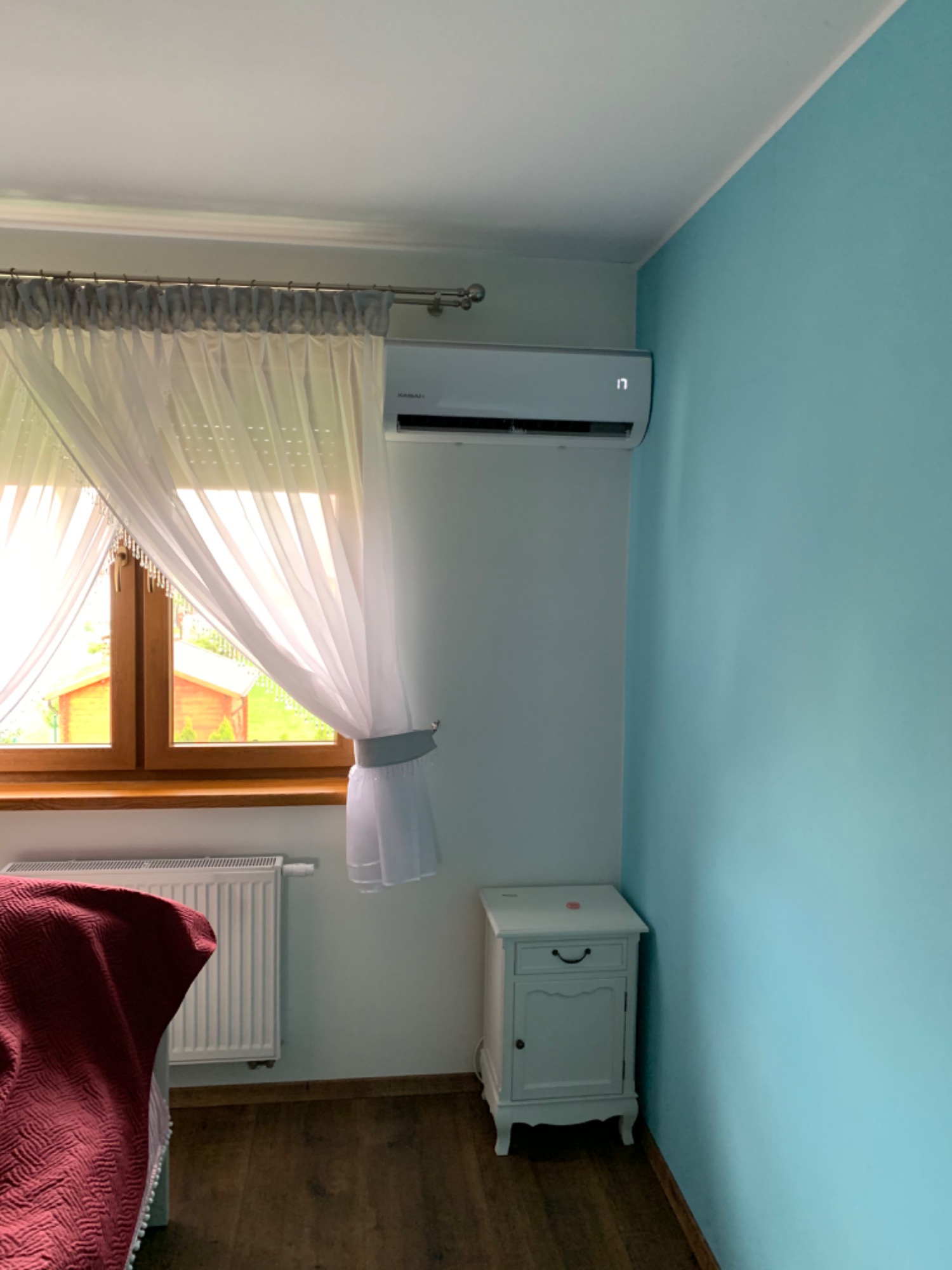 Biały klimatyzator ścienny zamontowany w pokoju z niebieską ścianą, obok okna z firankami i białej szafki nocnej.