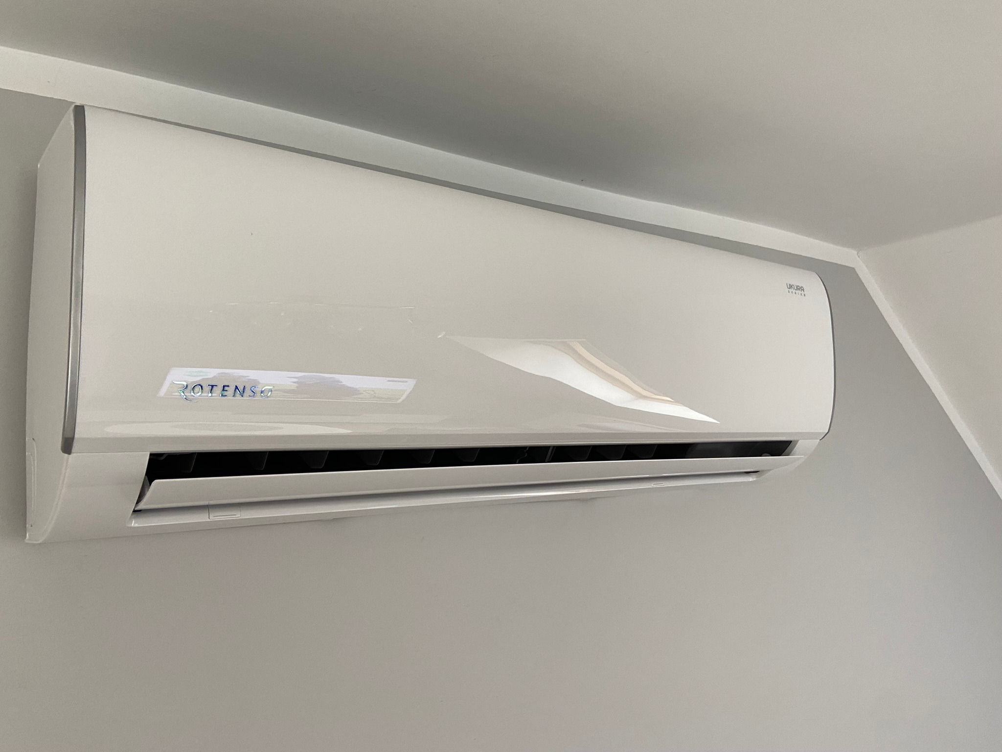 Biała jednostka wewnętrzna klimatyzacji Rotenso Ukura zamontowana na szarej ścianie, widoczny fragment sufitu w rogu.