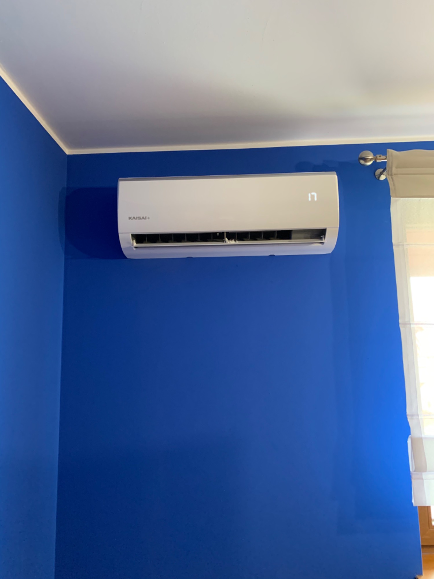 Biały klimatyzator Kaisai+ zamontowany na intensywnie niebieskiej ścianie, z widocznym fragmentem okna z roletą po prawej stronie.