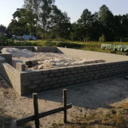 Wzniesione ściany fundamentowe z bloczków betonowych wypełnione piaskiem, widok na otoczenie z drzewami i częściowo widocznym budynkiem w tle.