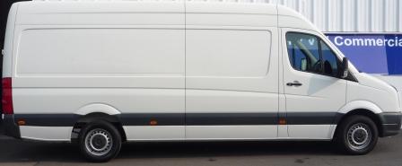 Biały, długi furgon dostawczy z ciemnoszarym pasem dolnym, zaparkowany przed budynkiem z blachy falistej, widziany z boku.