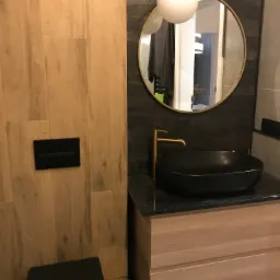 Nowoczesna łazienka z drewnianymi płytkami, czarną umywalką nablatową i okrągłym lustrem z podświetleniem kulistym.