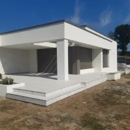 Nowoczesny dom z białymi ścianami i tarasem wyłożonym jasnoszarymi płytami, widoczne schody tarasowe, rośliny w donicach, z roletami zewnętrznymi w oknach, na tle błękitnego nieba.