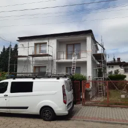 Dwupiętrowy dom w trakcie prac elewacyjnych, otoczony rusztowaniami, z białym busem firmy parkującym przed posesją.