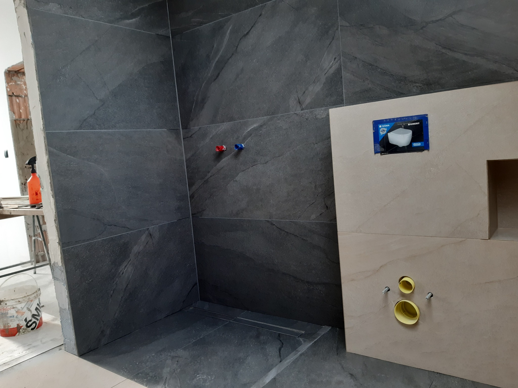 Częściowo wykończona łazienka w Nowym Sączu z ciemnymi płytkami imitującymi kamień na ścianach i podłodze, widoczny odpływ liniowy w strefie prysznica oraz zabudowa podtynkowego stelaża WC...