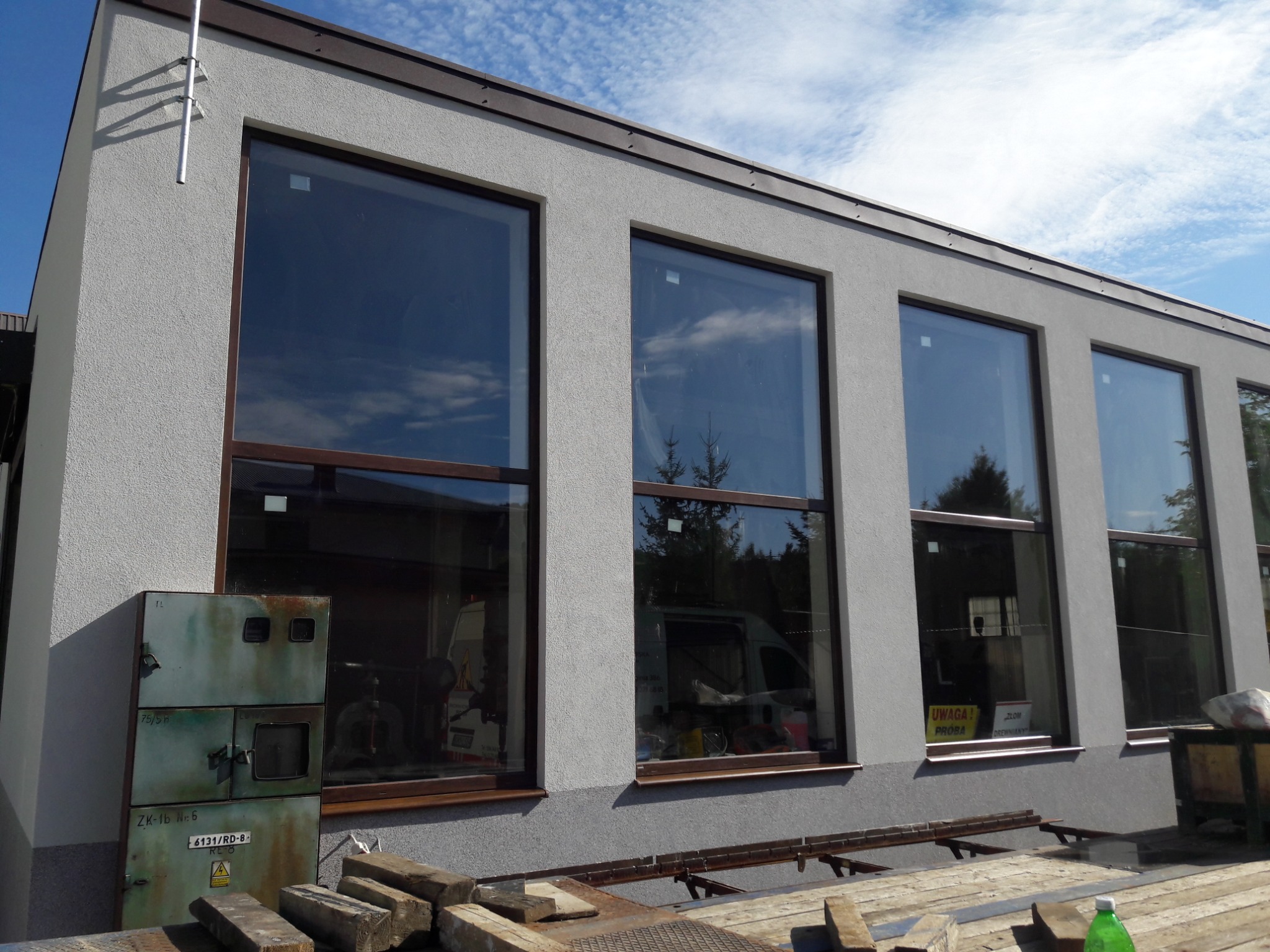 Elewacja budynku z dużymi oknami, szara struktura tynku, widoczne elementy rusztowania i skrzynka elektryczna z ostrzeżeniem, niebo z chmurami w tle.