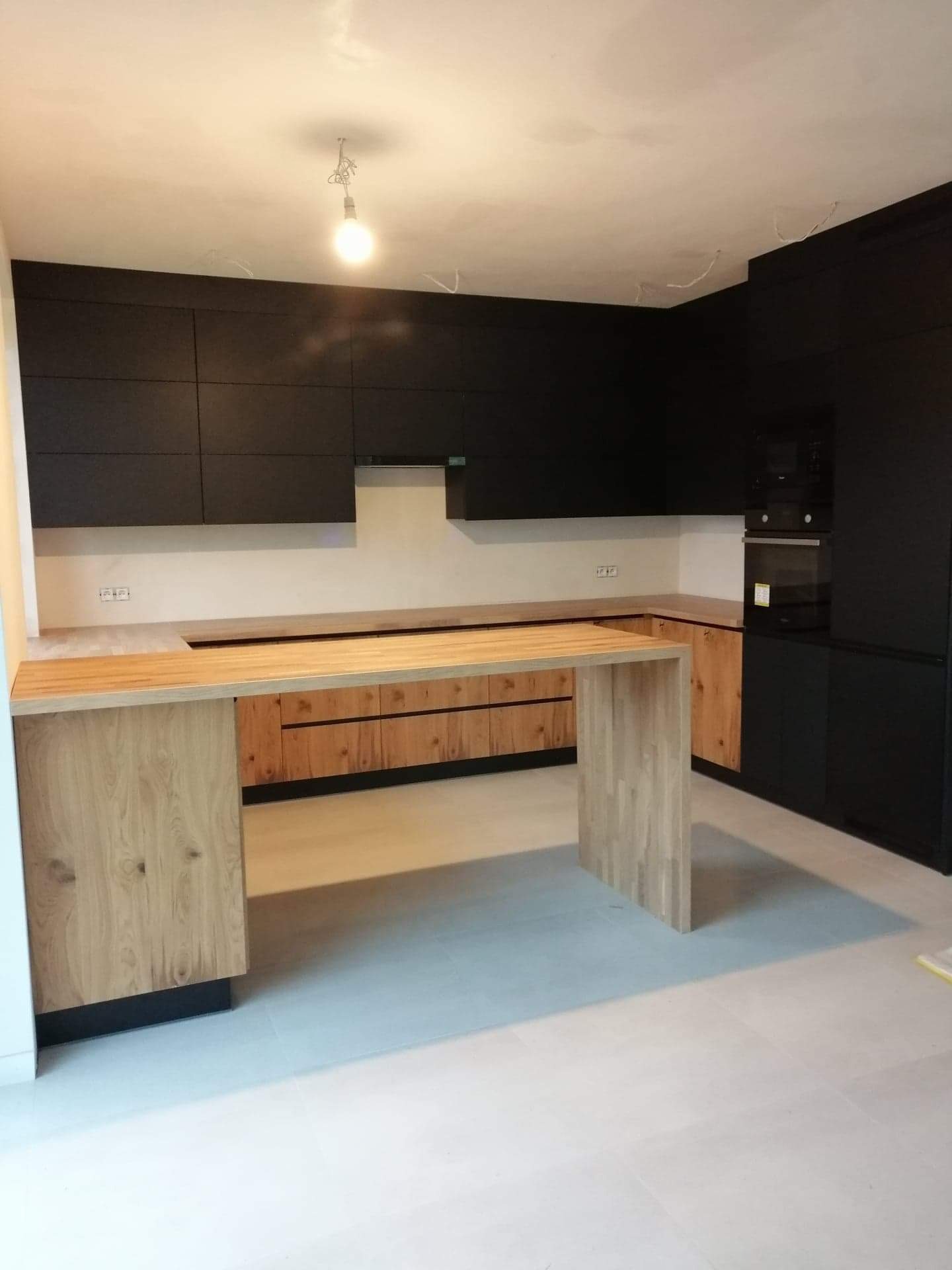 Nowoczesna kuchnia z czarnymi szafkami górnymi i drewnianymi dolnymi, wyspa z blatem z jasnego drewna, surowa żarówka zwisająca z sufitu, widoczne niedociągnięcia w wykończeniu sufitu.